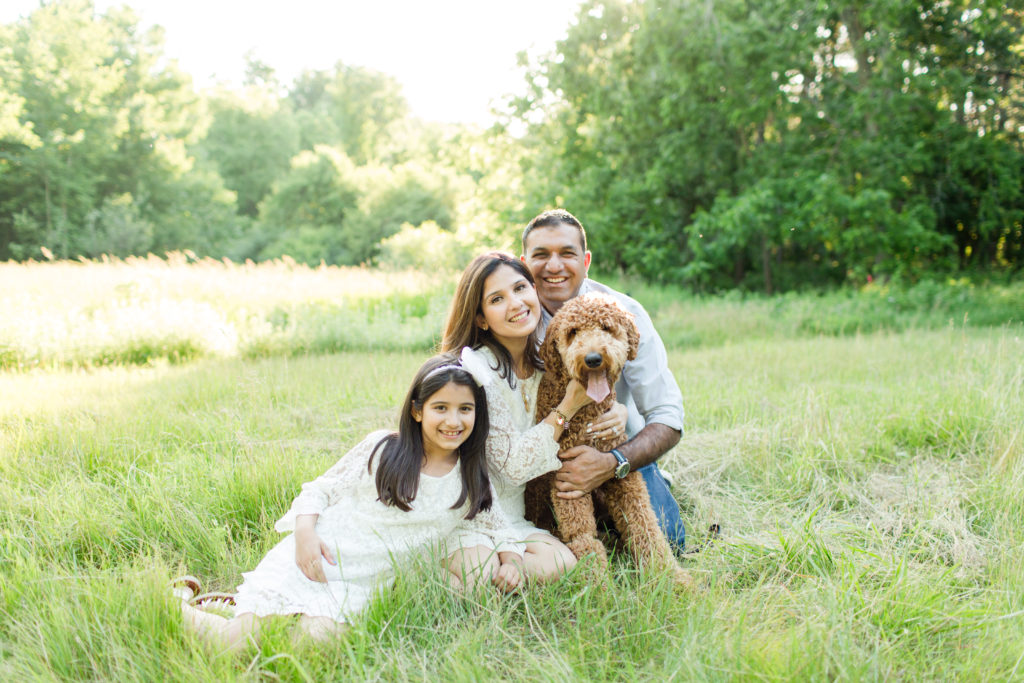 Holliston Family Photographer - Young family sitting in field with their fluffy dog, smiling for family photo session in Sherborn Massachusetts.