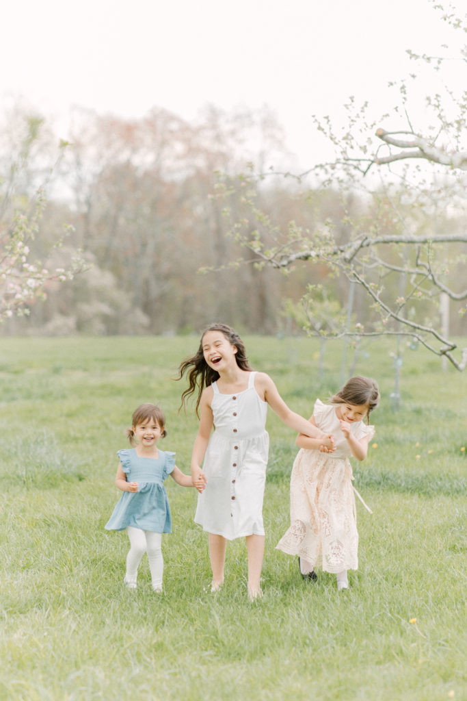 Three sisters holding hands and laughing during spring photo session with Boston family photographer Corinne Isabelle