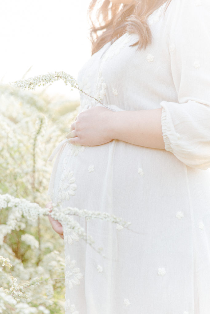 Close up photo of expecting mom's baby bump posing for maternity photos with Boston Maternity Photographer Corinne Isabelle Photography at the Arnold Arboretum in the spring with white floral blooms