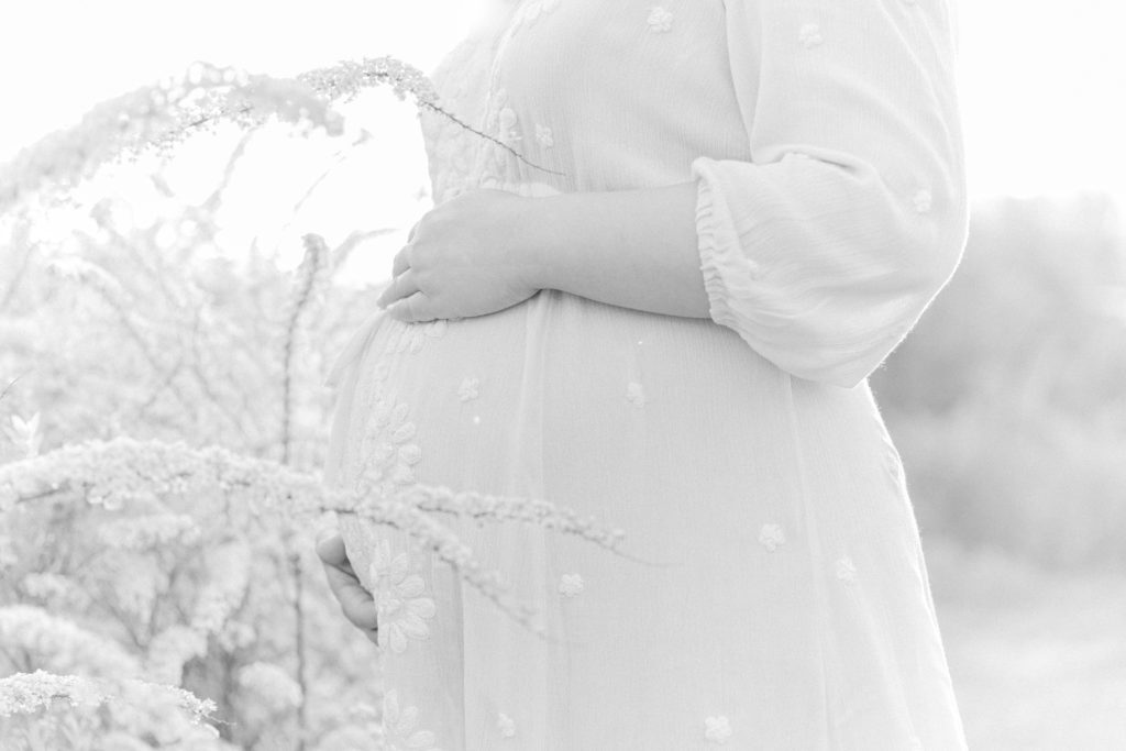 Black and white close up photo of expecting mom's baby bump posing for maternity photos with Boston Maternity Photographer Corinne Isabelle Photography at the Arnold Arboretum in the spring with white floral blooms