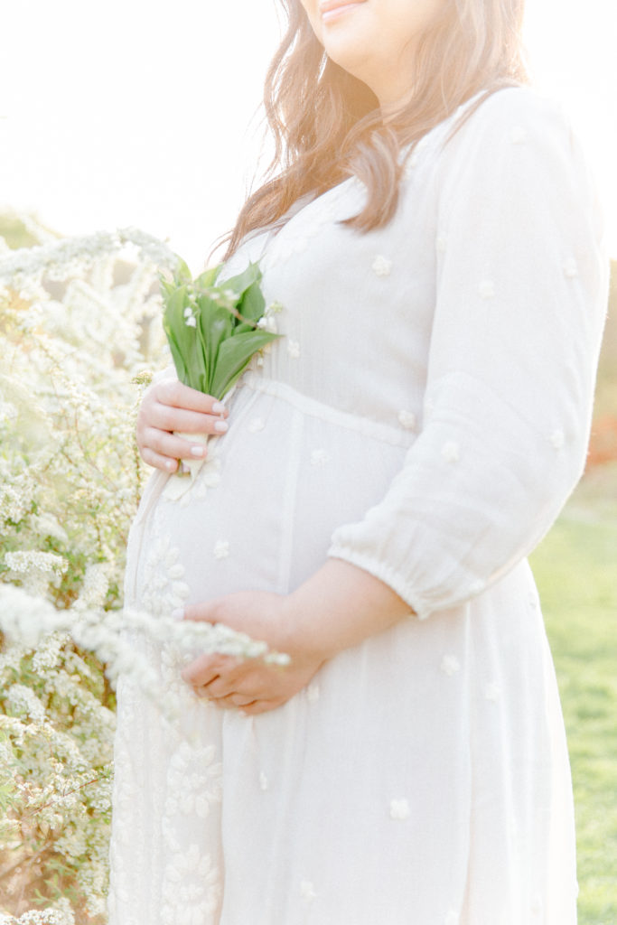 Close up photo of expecting mom's baby bump posing for maternity photos with Corinne Isabelle Photography at the Arnold Arboretum in the spring with white floral blooms