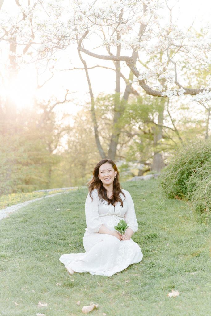 Photo of expecting mom sitting and posing for maternity photos with Boston Maternity Photographer Corinne Isabelle Photography at the Arnold Arboretum in the spring with white floral blooms