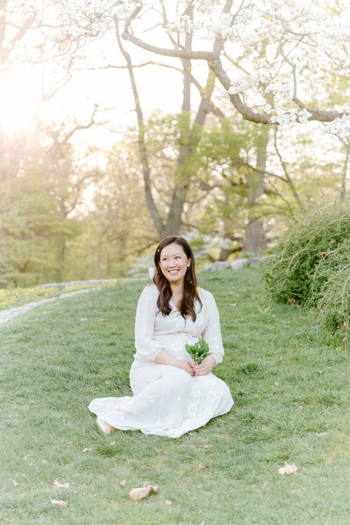 Photo of expecting mom sitting and posing for maternity photos with Corinne Isabelle Photography at the Arnold Arboretum in the spring with white floral blooms