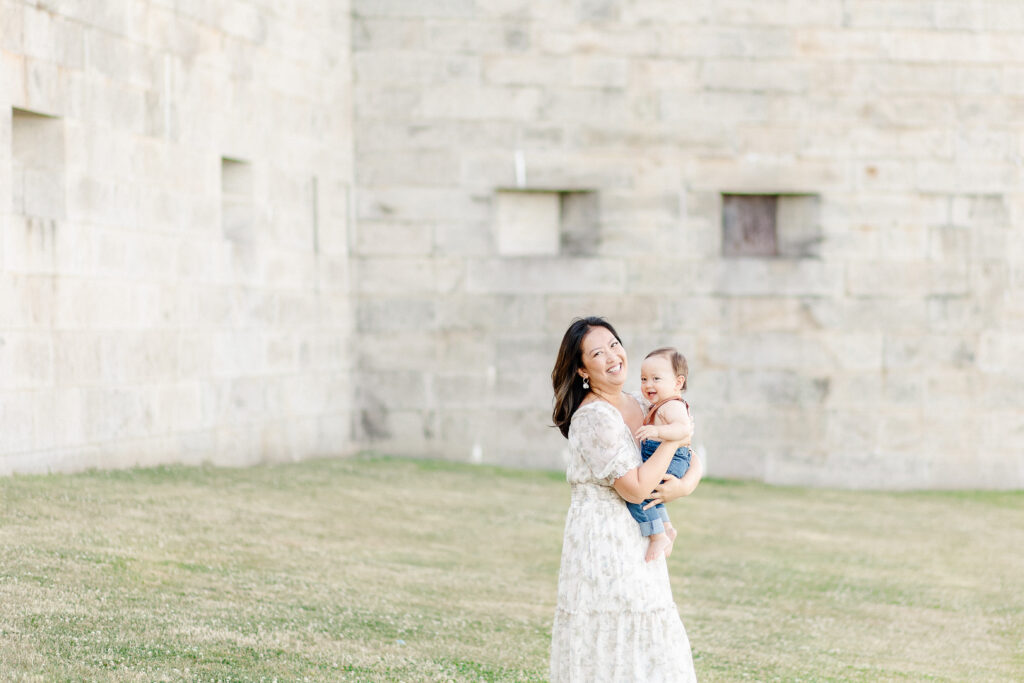 A mother wearing a light colored floral dress while holding her son and laughing towards the camera during photo session at Castle Island in Boston, MA with Corinne Isabelle Photography