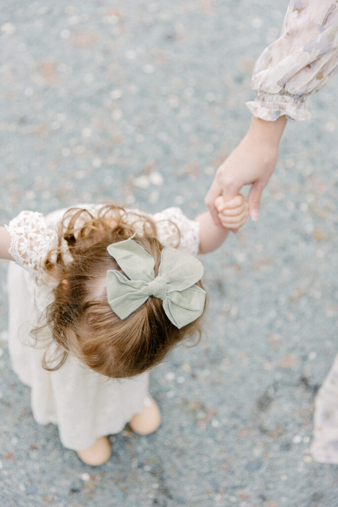 A young girl in a white dress standing on a gravel pathway while holding her mom's hand during photo session with Boston photographer Corinne Isabelle