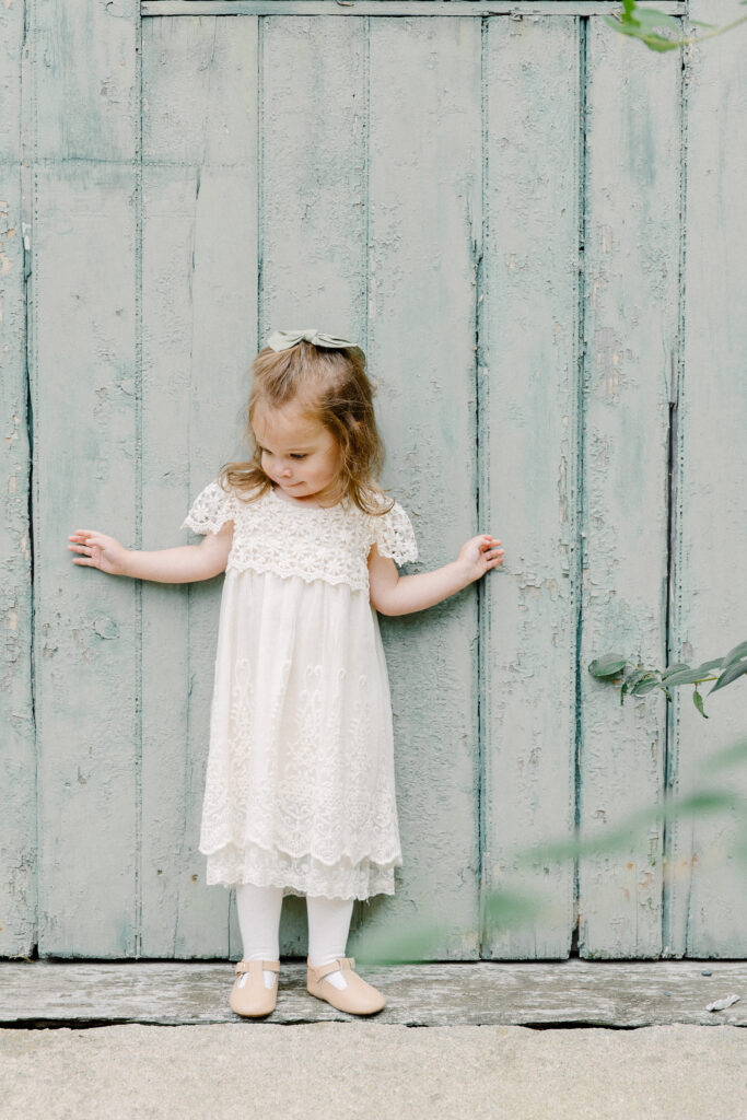 A young girl in a white dress standing in front of a mint green barn door during photo session with Boston photographer Corinne Isabelle